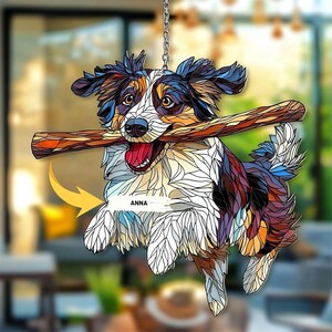 May include: A colorful, stained-glass style dog ornament with a stick in its mouth. The dog is depicted in shades of brown, blue, white, and black, with the name "ANNA" on the side. The ornament hangs from a silver chain.