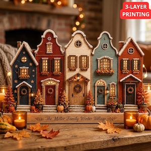 May include: A decorative wooden winter village display featuring five connected house structures in blue, red, cream, and green. The base is engraved with the text "The Miller Family Winter Village". It includes miniature pumpkins and trees.