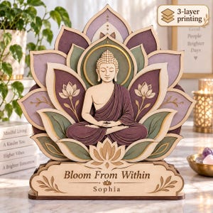 Könnte beinhalten: Ein mehrschichtiges Holzkunstwerk mit einer zentralen Buddha-Figur in Lotus-Sitzhaltung, umgeben von stilisierten Lotusblütenblättern in Violett-, Grün- und Cremetönen. Der Sockel zeigt den Text Bloom From Within und den Namen Sophia.