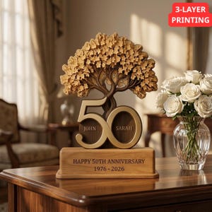 Op de afbeelding: Een houten 50-jarig jubileumdisplay met een lasergesneden boom, het getal 50 met de namen John en Sarah, en een voetstuk gegraveerd met HAPPY 50TH ANNIVERSARY 1976-2026. Een glazen vaas met witte rozen staat ernaast.