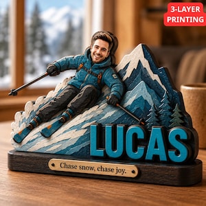 Peut inclure: Un présentoir en bois multicouche représentant un skieur en veste bleue et pantalon noir sur une montagne enneigée. Le nom LUCAS est écrit en grandes lettres bleues, et une plaque indique Chase snow, chase joy. Une étiquette rouge dit 3-LAYER PRINTING.