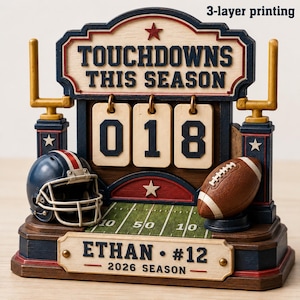 Puede incluir: Un marcador de fútbol americano decorativo con el texto TOUCHDOWNS THIS SEASON, una etiqueta 3-layer printing, un casco azul oscuro, un balón de fútbol americano marrón y una base con el texto ETHAN #12 2026 SEASON y un campo verde.
