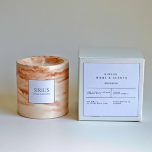 Pode incluir: Uma vela cilíndrica com um design de mármore e um rótulo branco que diz "SIRIUS HOME & SCENTS". Ao lado, uma caixa branca com a mesma marca e a palavra "BOURBON". A vela é feita com cera de soja natural.