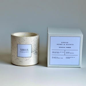 Pode incluir: Uma vela cilíndrica num suporte de betão salpicado e de cor clara, ao lado de uma caixa branca. A vela tem um rótulo branco que diz "SIRIUS HOME & SCENTS". A caixa tem um rótulo que diz "SIRIUS HOME & SCENTS SENSUAL AMBER".