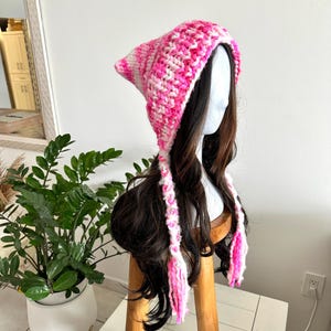 May include: A handmade crochet hat in shades of pink and white, featuring a pointed top and long, braided ties with pink tassels. The hat is displayed on a mannequin head with long, dark brown hair.