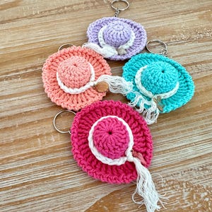 May include: Four crocheted hat keychains in different colors: lavender, coral, turquoise, and pink. Each keychain has a white tassel and a silver key ring.
