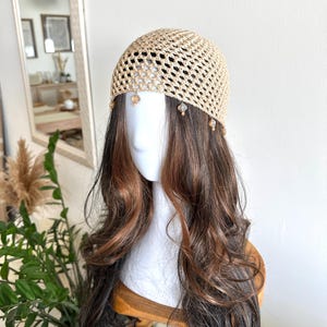 May include: A beige crocheted hat with a netted design, adorned with small, dangling amber-colored beads. The hat is displayed on a mannequin head with long, wavy brown hair. The background includes a mirror and greenery.
