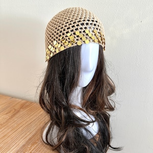 May include: A beige crocheted cap with a mesh design, adorned with a row of gold-colored circular sequins along the front. The cap is displayed on a mannequin head with long, wavy brown hair. The background is a plain, light-colored wall.