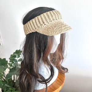 May include: A beige crochet visor with a textured, ribbed band and a firm, protruding brim. The accessory is crafted from yarn in a neutral tan shade, featuring a consistent stitch pattern throughout the crown and the front-facing sun visor.