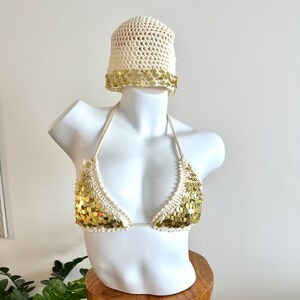 Pode incluir: Um top de biquíni de crochê creme e chapéu de balde a condizer adornados com lantejoulas douradas. O top do biquíni tem alças de cabresto e formato triangular. O chapéu tem uma aba larga com lantejoulas douradas. O conjunto é exibido em um manequim branco.