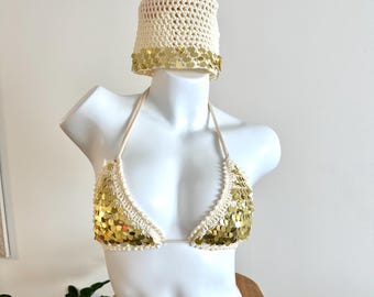 May include: A cream-coloured crochet bikini top and matching bucket hat decorated with gold sequins. The bikini top has halter straps and a triangle shape. The hat has a wide brim with gold sequins. The set is displayed on a white mannequin.