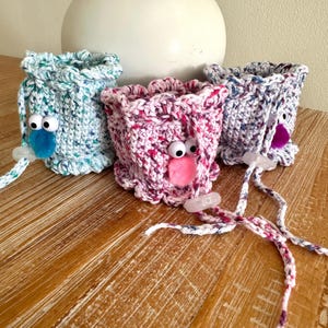 May include: Three crocheted cup cozies with whimsical faces. Each cozy features a different color pattern: blue, pink, and purple. The cozies have googly eyes and a pom-pom nose.