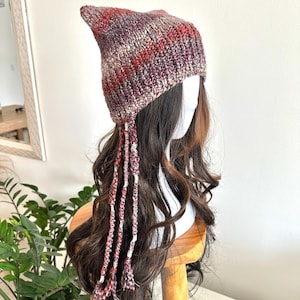 May include: A hand-knitted hat in shades of burgundy, red, and cream, featuring a pointed top and long, braided tassels. The hat is displayed on a mannequin head with long, wavy brown hair. The hat's texture is chunky and warm, perfect for cold weather.