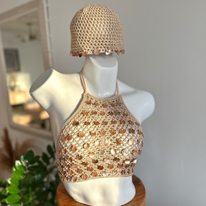 May include: A beige crochet halter top and matching beanie hat set. Both pieces are crafted from a metallic-toned yarn and are embellished with numerous small, circular, iridescent shell-like sequins that catch the light across the surface.