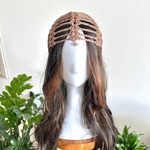 May include: A brown crocheted headpiece with a geometric design, adorned with a small, silver-toned charm featuring an eye design. The headpiece is displayed on a mannequin head with long, wavy brown hair with auburn highlights.