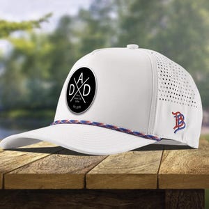 May include: White baseball cap with a black circular patch featuring the letters "DAD" in a cross design. The cap has a mesh back, a curved brim, and a blue and red rope detail. A small logo is embroidered on the side.