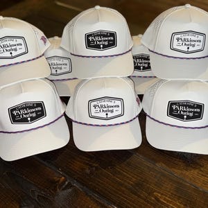 May include: A collection of white baseball caps with a mesh back and a curved brim. Each cap features a black and white patch with the text "CATHERINE'S Parkinson's Outing" and a decorative rope trim.
