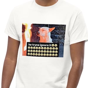 May include: A white t-shirt featuring a graphic print of a light-colored dog with eyes closed next to a flame. Below the dog, a black rectangular box contains the white text "he tryna ignore it" followed by rows of crying face emojis.