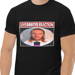 May include: A black short-sleeved t-shirt featuring a rectangular graphic print. The graphic contains the white text "LIVE GOATIS REACTION" on a red background above an oval image of a person with long hair wearing a white shirt and a microphone.