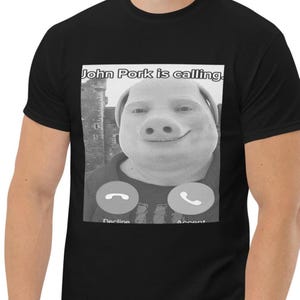Könnte beinhalten: Schwarzes T-Shirt mit einem Graustufenbild eines Schweinegesichts und dem Text "John Pork is calling." Darunter befinden sich zwei Telefonsymbole mit den Wörtern "Decline" und "Accept."