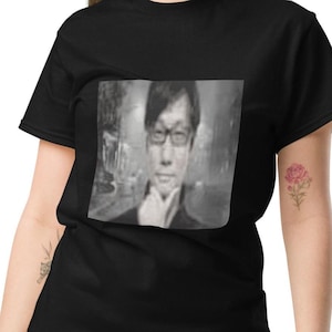 May include: Black t-shirt with a grayscale portrait of a person wearing glasses. The image shows a person with their hand near their chin. The t-shirt is made of a soft material.