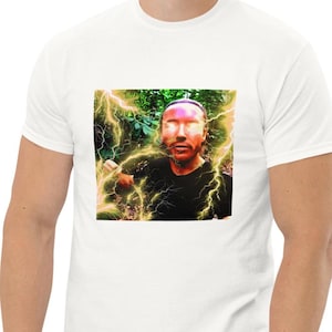 May include: A white short-sleeved t-shirt featuring a square graphic print on the chest. The image depicts a person with glowing eyes surrounded by yellow lightning bolts against a background of green foliage, wearing a black shirt.