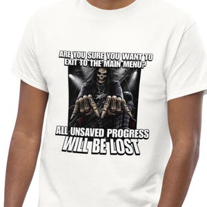 May include: A white t-shirt featuring a graphic of a skeleton musician with long dark hair and leather clothing. The text reads: ARE YOU SURE YOU WANT TO EXIT TO THE MAIN MENU? ALL UNSAVED PROGRESS WILL BE LOST. The skeleton has HARD ROCK on its knuckles.