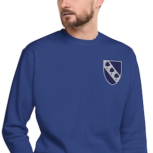 May include: A royal blue sweatshirt with a small embroidered crest on the chest. The crest is a navy blue shield with a silver border, featuring three silver trees arranged diagonally. The sweatshirt has a crew neck and long sleeves.