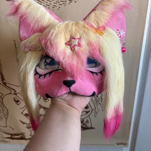 May include: A pink and yellow furry fox mask with large ears, a black nose, and blue eyes. The mask is decorated with a star, a butterfly, and small pink flowers. The mask is held up by a hand.