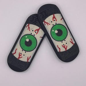 Puede incluir: Dos fingerboards negros con fondo blanco y un gráfico de ojo verde. El ojo tiene venas rojas y una pupila negra.