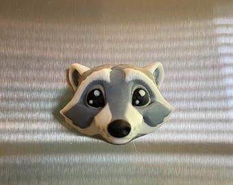 May include: A close-up of a gray and white raccoon face figurine with black eyes and a black nose. The figurine is made of a smooth, glossy material.