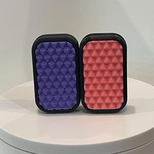 Pode incluir: Dois objetos pequenos, retangulares, com moldura preta e superfícies texturizadas. Uma superfície é roxa e a outra é rosa.