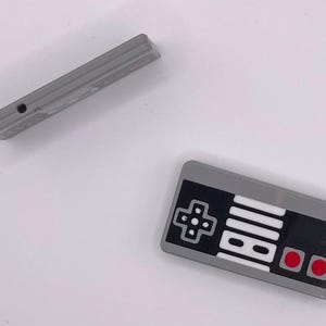 May include: A grey and black Nintendo Entertainment System controller with red buttons. The controller is made of plastic and has a classic design.