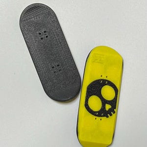 Puede incluir: Dos fingerboards, uno negro y otro amarillo con un gráfico de calavera negra.