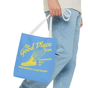 Pode incluir: Uma bolsa tote azul com alças brancas. A bolsa tem um gráfico amarelo de um sapato alado com o texto "The Good Place Farm" e "Chattahoochee Hills, GA. Where heaven is a place on earth."