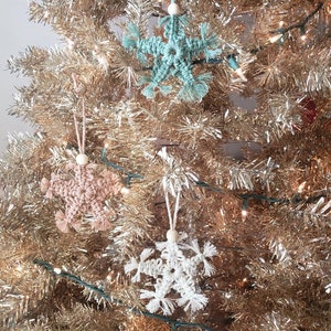 May include: Three crocheted snowflake ornaments hanging on a gold tinsel Christmas tree. The snowflakes are in shades of teal, beige, and white.