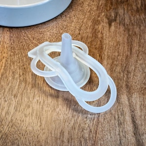 May include: A clear, soft plastic bottle nipple with a looped handle design. The nipple has a rounded base and a narrow, elongated tip. The design is made of a flexible, translucent material. The background is a wooden surface.
