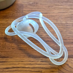 Può includere: Una scultura astratta traslucida con una forma centrale arrotondata e linee ad anello interconnesse. La scultura è realizzata in un materiale di colore chiaro e poggia su una superficie di legno. Il design è intricato e sembra essere un oggetto decorativo.