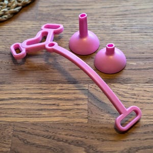 May include: A pink plastic accessory set consisting of a long, curved handle with rectangular openings at both ends and two separate pink conical suction cup attachments. The components are smooth, solid-colored, and feature a simple, functional design.