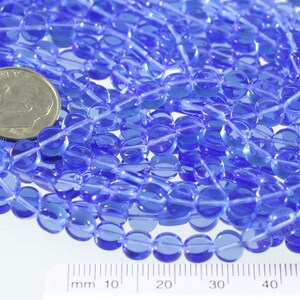 May include: A close-up of a pile of translucent blue glass beads. The beads are round and have a slightly oval shape. They are approximately 6mm in diameter. A ruler is visible in the bottom of the image, showing the size of the beads.