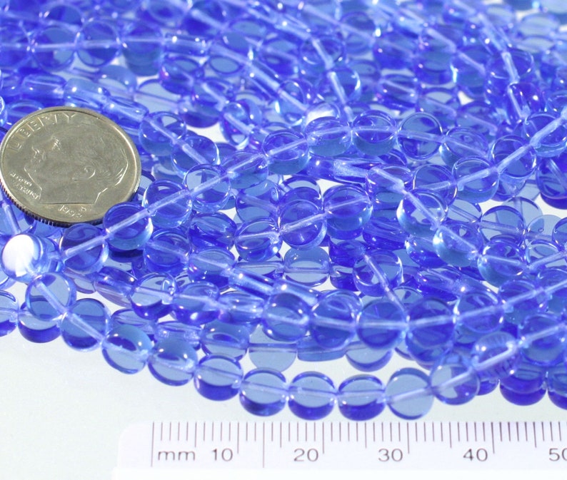 May include: A close-up of a pile of translucent blue glass beads. The beads are round and have a slightly oval shape. They are approximately 6mm in diameter. A ruler is visible in the bottom of the image, showing the size of the beads.