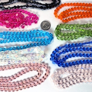 May include: Assortment of round glass beads in various colors, including pink, blue, green, orange, white, and black. The beads are arranged in rows on a white surface.