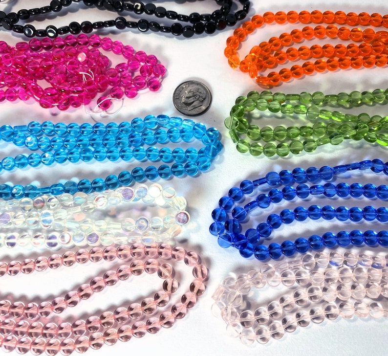 May include: Assortment of round glass beads in various colors, including pink, blue, green, orange, white, and black. The beads are arranged in rows on a white surface.