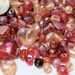 May include: Assortment of red, pink, and clear glass beads in various shapes, including hearts, rounds, and ovals. The beads have a shiny, iridescent finish.