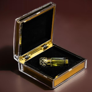 May include: A clear glass bottle with a gold cap and a gold label inside a clear plastic box with a gold trim. The bottle contains a clear liquid.