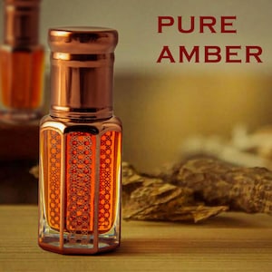 May include: A small bottle of amber-coloured perfume with a copper-coloured cap and a decorative pattern on the bottle. The bottle is sitting on a wooden surface with a piece of wood behind it. The text "PURE AMBER" is written in red letters on a white background.