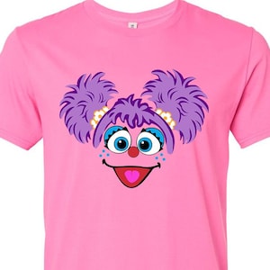 May include: Pink t-shirt featuring a cartoon character's face. The character has purple hair, blue eyes, and a red tongue. The design is centered on the shirt, creating a playful and colorful look. The shirt is made of a soft material.