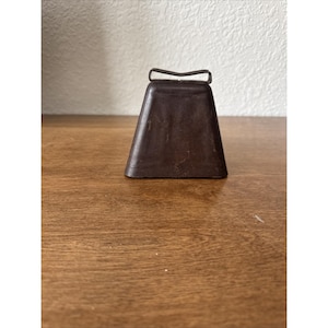 May include: A dark brown, trapezoidal metal cowbell with a weathered, matte finish. The bell features a simple, rectangular metal loop handle attached to the top center, showcasing a utilitarian design with visible signs of age and use.