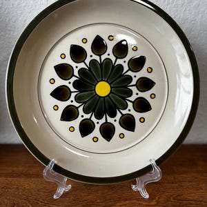 May include: A round ceramic plate with a cream-colored base and a dark green rim. The center features a stylized floral pattern with dark green petals surrounding a bright yellow circular core, accented by small yellow dots in a circle.