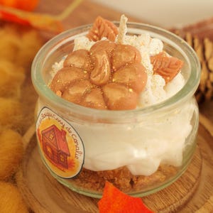 Peut inclure: Une bougie décorative dans un bocal en verre transparent, avec un design en couches comprenant une base de biscuit, de la crème blanche et des éléments décoratifs bruns. La bougie a une mèche et est posée sur une surface en bois. Un autocollant avec une illustration de maison est sur le bocal.